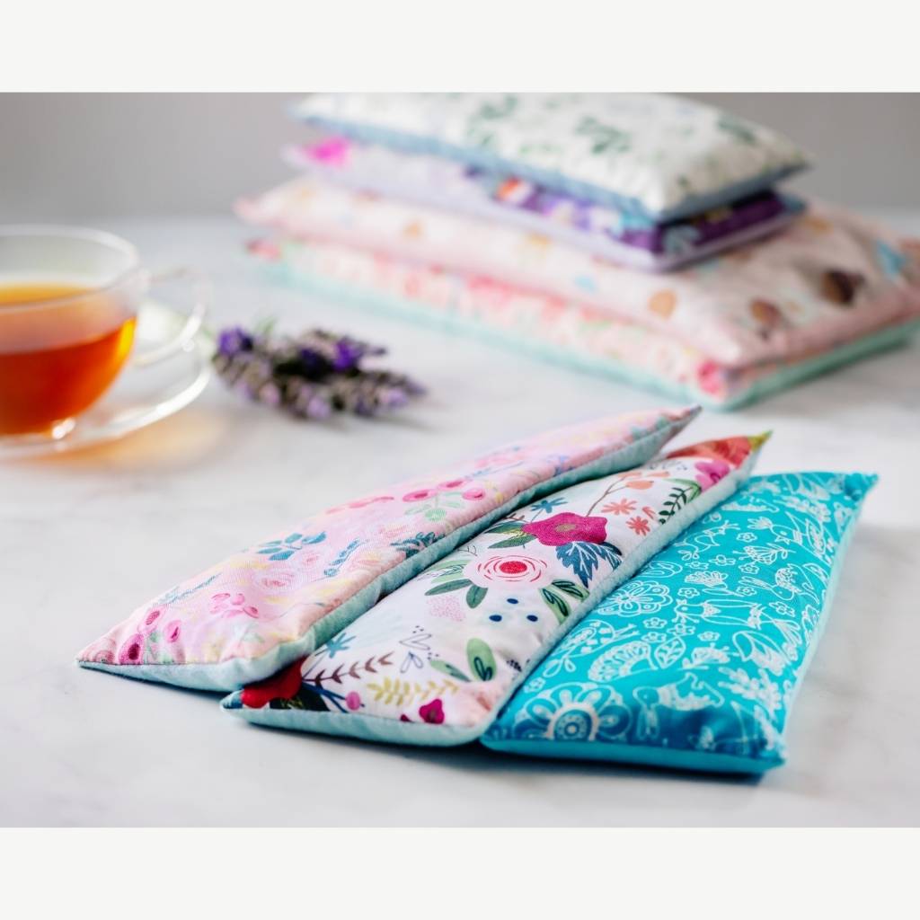 Three microwavable heating pads laid out in different floral fabrics and a stack of heating pads next to a sprig of lavender.